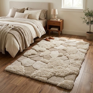 May include: A rectangular area rug with a stone-like pattern in shades of beige and cream. The rug is placed on a hardwood floor in a bedroom setting, near a bed with neutral-colored bedding. The rug's design adds a textured, natural element to the room.