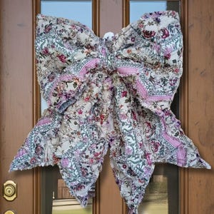 May include: A large, decorative bow made of floral fabric, featuring a cream background with pink, purple, and green floral patterns. The bow is attached to a wooden door. The bow's dimensions are approximately 76 cm wide, creating a welcoming aesthetic.