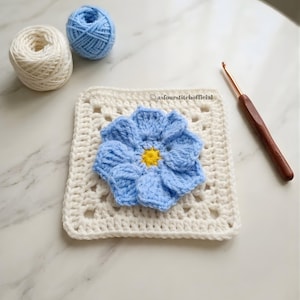 Op de afbeelding: Een wit gehaakt vierkant in granny square-stijl met een lichtblauwe bloem met een geel hart in het midden. De tekst asfourstitchofficial staat boven de bloem. Twee bollen garen in wit en lichtblauw en een houten haaknaald zijn aanwezig.