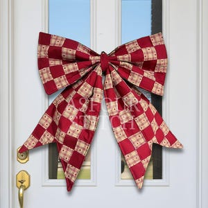 May include: A large decorative bow featuring a red and cream checkered pattern with small heart motifs within the squares. The fabric bow has two wide top loops and two long, pointed tails hanging downwards, centered on a white door.