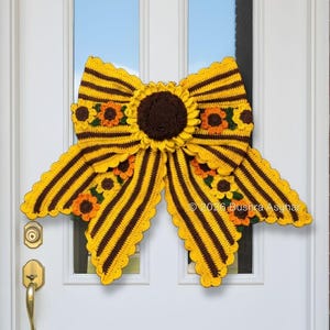 May include: A handmade crochet bow in yellow and brown, resembling a sunflower. The bow is adorned with smaller sunflower accents and is displayed on a white door. The design includes scalloped edges and a central sunflower.