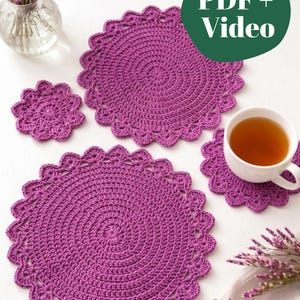 May include: A set of purple crocheted circular mats with scalloped edges, including two large mats and two smaller coasters. One coaster supports a white ceramic cup filled with tea. A green circular graphic displays the text PDF + Video.