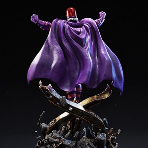 Op de afbeelding: Een beeld van een personage in een paarse cape en helm, armen omhoog. De figuur staat op een decoratieve basis met een donkere achtergrond. De cape is dieppaars en de helm is rood.