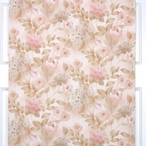 Puede incluir: Un rollo de papel pintado vintage con un patr&oacute;n floral repetitivo. El dise&ntilde;o incluye flores rosas y blancas, hojas y tallos marrones, sobre un fondo beige claro. El papel pintado se muestra contra un marco de ventana blanco.