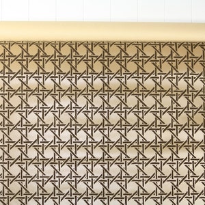 May include: A roll of tan-colored wallpaper with a repeating dark brown woven cane pattern. The wallpaper is partially unrolled, revealing the intricate design. The background is a white wooden surface.