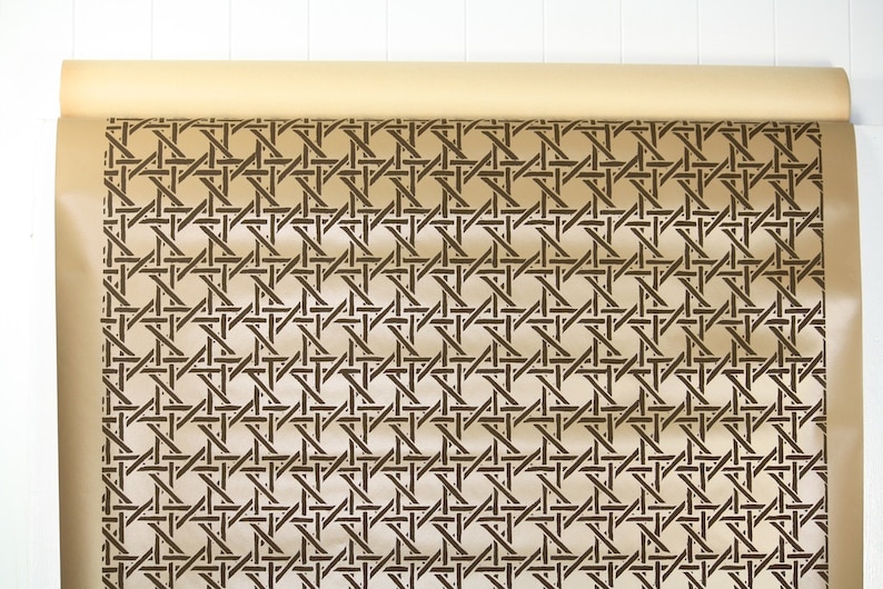 May include: A roll of tan-colored wallpaper with a repeating dark brown woven cane pattern. The wallpaper is partially unrolled, revealing the intricate design. The background is a white wooden surface.