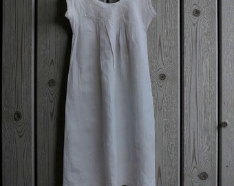 May include: A white nightdress with a scoop neck and cap sleeves. The dress has lace detailing around the neckline and sleeves, with vertical pleats down the front. It hangs on a wooden hanger against a wooden plank background. The dress is approximately 100 cm in length.