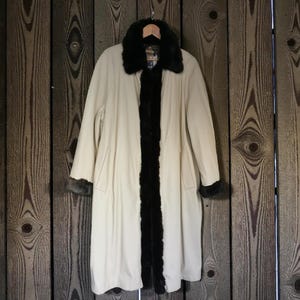 May include: A cream-colored coat with a dark fur collar and trim, hanging against a wooden background. The coat has long sleeves and a classic design, with a dark fur accent down the front.