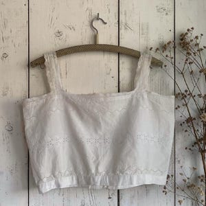 Op de afbeelding: Een witte mouwloze crop top met gedetailleerde broderie anglaise op de voorzijde en langs de brede schouderbanden. Het kledingstuk heeft een vierkante halslijn en een rechte zoom, met een verfijnd geborduurd patroon over de gehele stof.