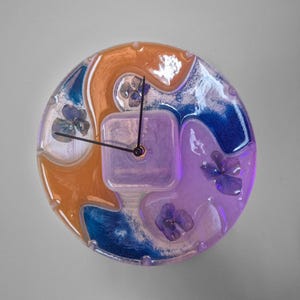 Puede incluir: Un reloj de pared redondo con un diseño de resina. La esfera del reloj presenta tonos naranja, azul y morado con flores secas incrustadas. Las manecillas del reloj son negras y el centro tiene forma cuadrada. Una pieza decorativa única.