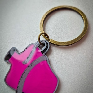 May include: A hot pink keychain with a gray outline and a brass-colored ring. The keychain is shaped like a coat or jacket. The background is a light, neutral color.