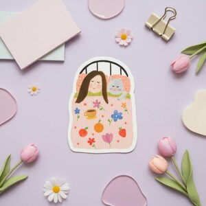 May include: A sticker depicting a woman and a grey cat in bed, under a pink blanket with flowers, fruit, and a coffee cup. The sticker is surrounded by pink flowers, tulips, and stationery on a lilac background.
