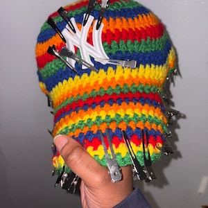 May include: A handmade, rainbow-striped crochet hat with a white New York Yankees logo. The hat is decorated with numerous silver hair clips around the brim. The hat is held by a person.