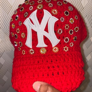 May include: A red, crocheted baseball cap with a white "NY" logo. The cap is embellished with numerous sparkling rhinestones in various colors. The hat is held up by a hand.