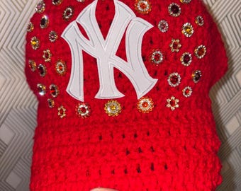 May include: A bright red crocheted baseball cap featuring a white embroidered New York Yankees logo on the front. The cap is decorated with numerous small, round, multi-colored rhinestones and flower-shaped crystals scattered across the crown.