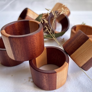 May include: A selection of wooden napkin rings, each crafted with a two-tone design. The rings are cylindrical, featuring sections of dark and light wood. They are displayed on a white surface, with some dried grass sprigs.