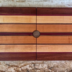 Puede incluir: Una tabla de cortar rectangular de madera con una variedad de tonos, incluyendo rayas marrón oscuro, marrón claro y púrpura rojizo. La tabla está dividida en dos secciones y tiene un asa circular en el centro.
