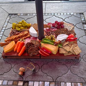 Puede incluir: Una tabla de embutidos de madera con una variedad de alimentos, incluyendo queso, galletas, uvas, aceitunas y embutidos. La tabla está dispuesta sobre una mesa exterior estampada. Un corcho de vino y un reloj de arena están en primer plano.