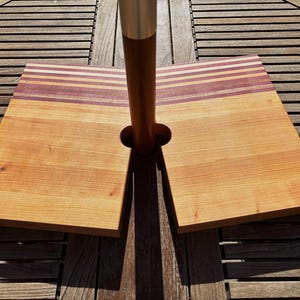 May include: A wooden umbrella base with two rectangular sections. The base is made of light-colored wood with a strip of darker wood and purple stripes. A brown pole with a silver top is centered between the two sections.
