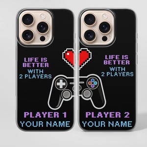 Può includere: Due custodie nere per telefono a tema gaming. Ogni custodia presenta un cuore rosso pixelato, una grafica del controller bianca e la scritta "LIFE IS BETTER WITH 2 PLAYERS". Sotto, c'è scritto "PLAYER 1 YOUR NAME" e "PLAYER 2 YOUR NAME" in viola.