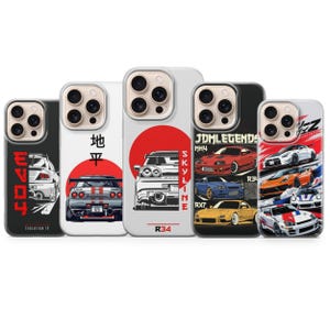 May include: A collection of five phone cases featuring car designs. The cases display various car models with graphics, including a red sun, Japanese text, and the words "JDM Legends." The cases are in black, white, and clear.