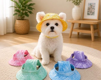 May include: A small white dog wearing a yellow tie-dye bucket hat with ear holes. Four additional bucket hats in pink, green, blue, and purple tie-dye patterns are arranged on the floor in front of the dog, each featuring a wide brim and strap.