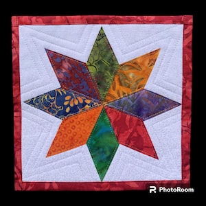 May include: A colorful, handmade star quilt block. The star is composed of diamond-shaped fabric pieces in various colors, including blue, orange, green, and red. The star is centered on a white background with a quilted pattern and a red border.
