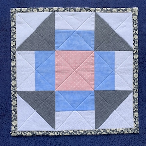 May include: A square fabric quilt block featuring a central pink square with white polka dots, surrounded by light blue and dark gray geometric shapes. The block is finished with a dark floral patterned border and visible diagonal stitching.