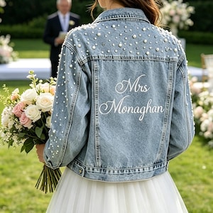 May include: Light blue denim jacket embellished with scattered pearls. The back of the jacket features the name "Mrs Monaghan" in elegant script. The jacket is worn over a white tulle skirt, with a bouquet of flowers in the foreground.