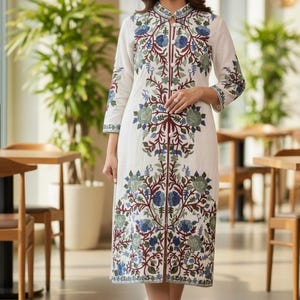 May include: A white, knee-length dress with floral embroidery in blue, green, and maroon. The dress features a mandarin collar and three-quarter sleeves. The dress is worn by a person in a well-lit indoor setting.