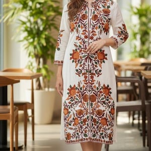May include: A cream-coloured, knee-length dress with detailed floral embroidery in orange, brown, and green. The dress has a mandarin collar, three-quarter sleeves, and a central zip. The dress is worn with brown sandals.