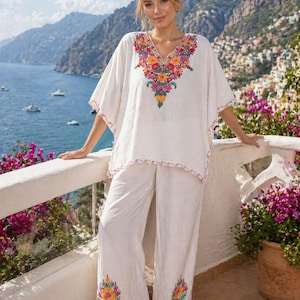 May include: A white linen two-piece outfit featuring a tunic top and wide-leg pants. The top has a V-neck and colorful floral embroidery. The pants have matching embroidery at the hem. The outfit is worn on a balcony overlooking a coastal landscape.