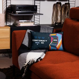 May include: Two decorative throw pillows with retro video game designs. One pillow features the text "CONTINUE? YES NO" with hearts. The other pillow has a colorful arcade machine graphic with "GAME OVER" text. The pillows rest on a rust-colored couch.