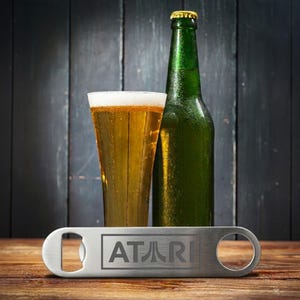 May include: A green beer bottle and a glass of beer with foam sit behind a silver bottle opener. The bottle opener has the Atari logo engraved on it. The items are on a wooden surface.
