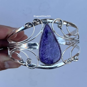 Pode incluir: Uma pulseira de punho de prata com uma grande pedra preciosa roxa em forma de gota. A pulseira tem um design aberto com redemoinhos decorativos e o texto "BhagwatiSilver925".