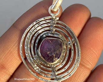 May include: A silver pendant with a raw, deep purple gemstone at its center. The pendant has a circular design with concentric, textured lines. The bail is a simple loop. The brand name "BhagwatiSilver925" is visible on the bottom left.