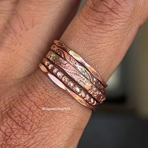 May include: A set of copper rings with detailed etched patterns, including floral and geometric designs. The rings are worn on a finger, highlighting the craftsmanship and the warm, reddish-brown colour of the metal. The rings are a unique piece of jewellery.