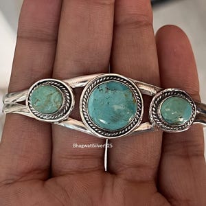 May include: A silver cuff bracelet featuring three round turquoise stones set in twisted rope borders. The central stone is larger than the two flanking stones. The metal band has a split design and the text BhagwatiSilver925 is visible.