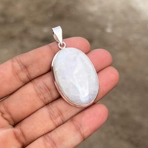May include: An oval-shaped moonstone pendant set in a polished silver frame. The gemstone features a smooth, translucent white surface with subtle iridescent reflections. A small silver bail is attached to the top for hanging on a chain.