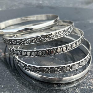 May include: A collection of silver bangles with various designs. Some bangles are plain, while others feature intricate patterns of flowers, circles, and geometric shapes. The bangles are stacked on a dark, textured surface.