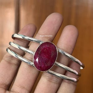 May include: A silver cuff bracelet featuring three parallel bands and a large, oval-shaped, faceted deep red ruby gemstone set in the center. The text BhagwatiSilver925 is visible on the metal surface near the gemstone setting.