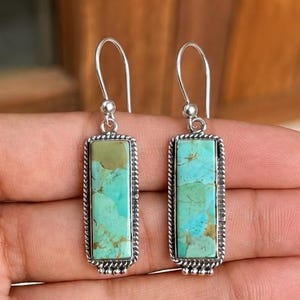 Puede incluir: Un par de pendientes colgantes con piedras turquesa rectangulares engastadas en marcos de plata con un detalle de borde de cuerda trenzada. Los pendientes cuelgan de ganchos de plata simples y muestran vetas naturales.