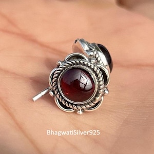 Może przedstawiać: Srebrne kolczyki sztyfty z okrągłym, głęboko czerwonym kamieniem szlachetnym. Kolczyki mają ozdobną, sznurowatą obwódkę i mały słupek. Widoczny jest tekst "BhagwatiSilver925".