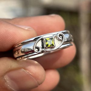 May include: A silver spinner ring featuring a central square-cut lime green gemstone. The band is accented with decorative silver scrollwork on either side of the stone, creating a detailed metal frame around the vibrant green square setting.