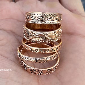 Könnte beinhalten: Fünf roségoldfarbene Ringe mit detaillierten floralen und geometrischen Mustern, gestapelt auf einer Hand. Jeder Ring weist aufwendige Designs auf. Die Ringe werden auf einer offenen Handfläche präsentiert, um die Details hervorzuheben. Die Farbe ist warm und metallisch.