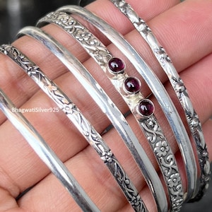 May include: A set of five silver bangles, including one plain band, two with intricate floral engravings, one with a smooth surface, and one featuring three small, round, deep red garnet gemstones set in a row along the metal band.