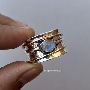 Op de afbeelding: Een zilveren spinnerring met een centraal ovaalvormig, lichtblauwe edelsteen. De ring heeft banden van zilver, koper en goudkleurig metaal. De ring wordt vastgehouden door de vingers van een persoon. De tekst "BhagwatiSilver925" is zichtbaar.