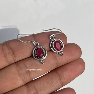 May include: A pair of silver drop earrings featuring oval-cut, deep red gemstones. Each earring has a circular frame around the gemstone and decorative accents at the bottom. The earrings are displayed against a light background.