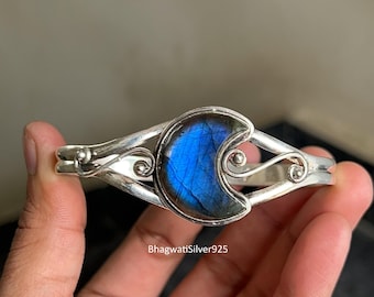 Puede incluir: Una pulsera de plata con una piedra de labradorita en forma de media luna, con destellos azules e iridiscentes. La pulsera tiene un diseño calado con detalles de volutas. El texto "BhagwatiSilver925" es visible.