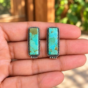 May include: A pair of rectangular stud earrings featuring vibrant turquoise stones with natural brown matrix patterns. Each stone is set in a polished silver frame with a twisted rope border and a small beaded detail at the bottom edge.